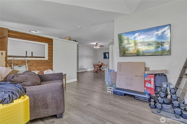 living room featuring ceiling fan, wood walls, baseboards, and wood finished floors