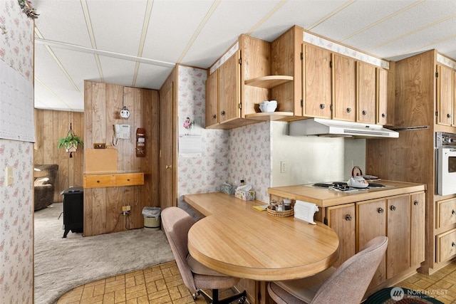 kitchen featuring stovetop, under cabinet range hood, wallpapered walls, wall oven, and brick patterned floors