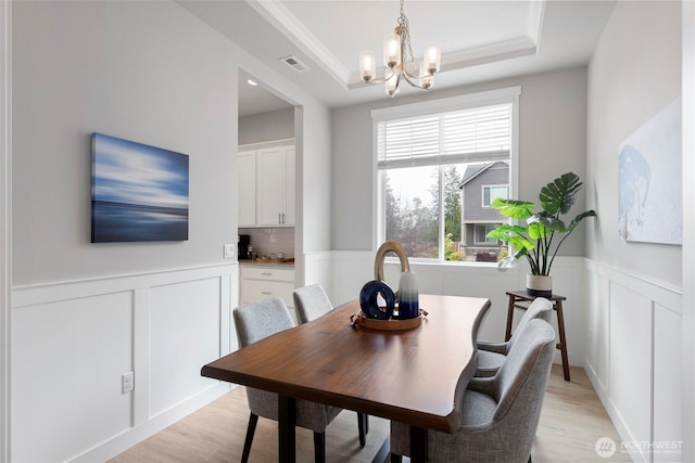 dining space with a decorative wall, a chandelier, light wood-style flooring, a tray ceiling, and a wainscoted wall