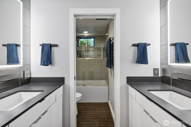full bath featuring two vanities, wood finished floors, toilet, and enclosed tub / shower combo