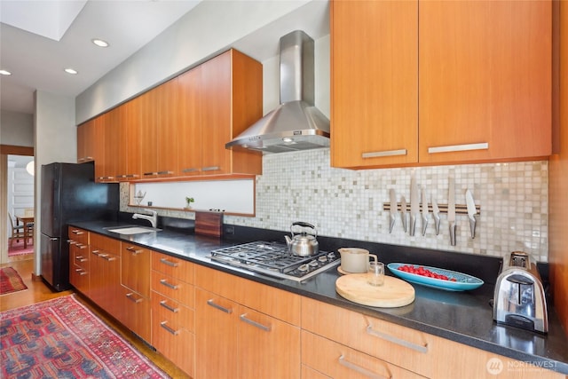 kitchen with wall chimney exhaust hood, stainless steel gas stovetop, a sink, tasteful backsplash, and freestanding refrigerator