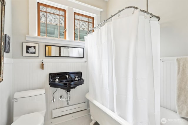 bathroom with a baseboard heating unit, wainscoting, toilet, shower / bath combo, and a decorative wall