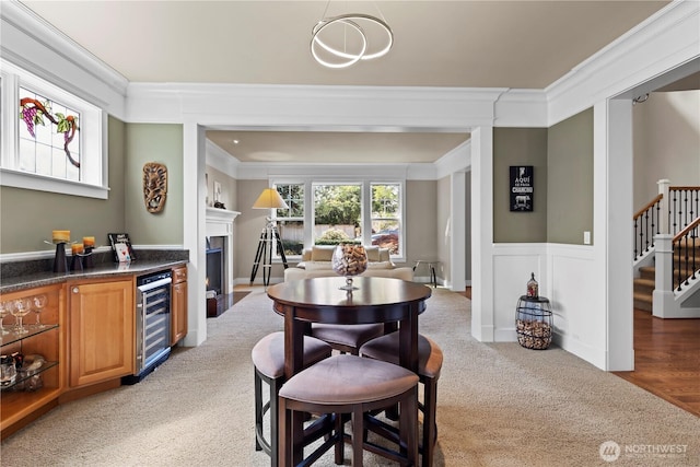 dining space featuring beverage cooler, stairway, ornamental molding, a fireplace with flush hearth, and light carpet