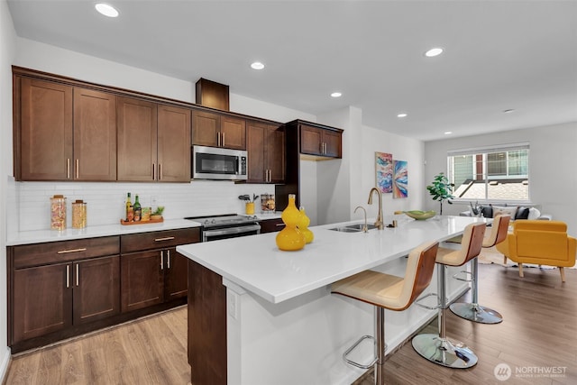 kitchen with appliances with stainless steel finishes, a sink, a center island with sink, light wood-type flooring, and light countertops