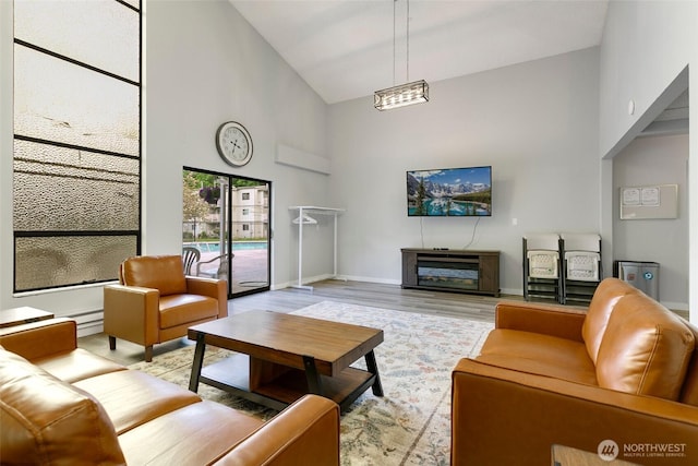 living room featuring high vaulted ceiling, light wood-style flooring, baseboard heating, baseboards, and a glass covered fireplace