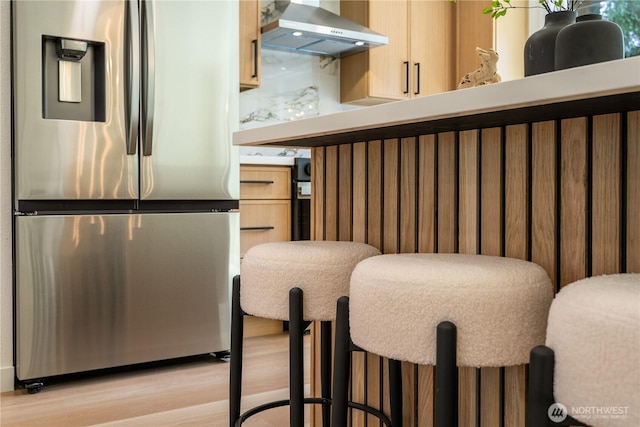 kitchen featuring stainless steel refrigerator with ice dispenser, wall chimney range hood, light wood-style flooring, and light brown cabinets