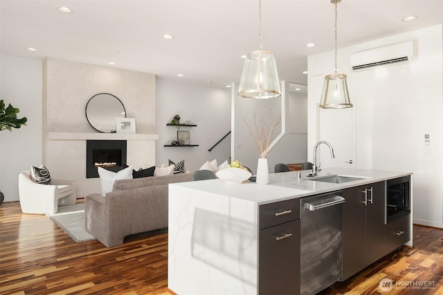 kitchen featuring an island with sink, a wall unit AC, a sink, stainless steel dishwasher, and dark wood-style flooring