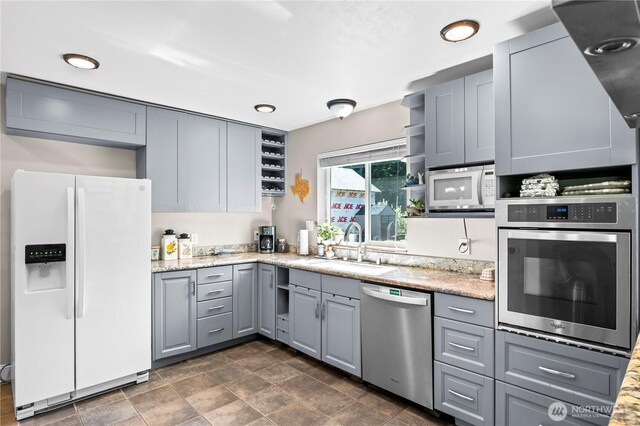 kitchen featuring stainless steel appliances, open shelves, gray cabinets, a sink, and light stone counters