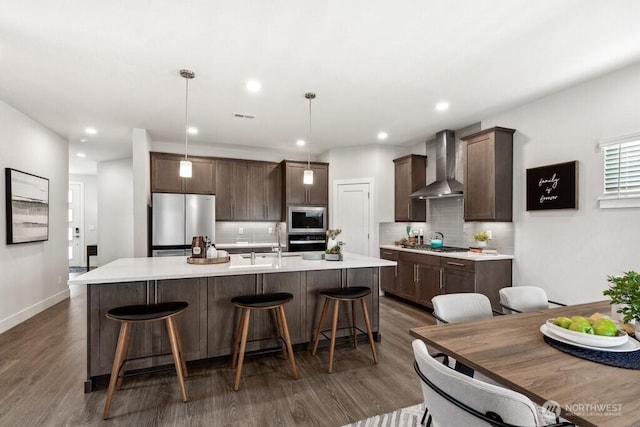 kitchen with dark brown cabinets, decorative backsplash, wall chimney exhaust hood, appliances with stainless steel finishes, and dark wood-style floors