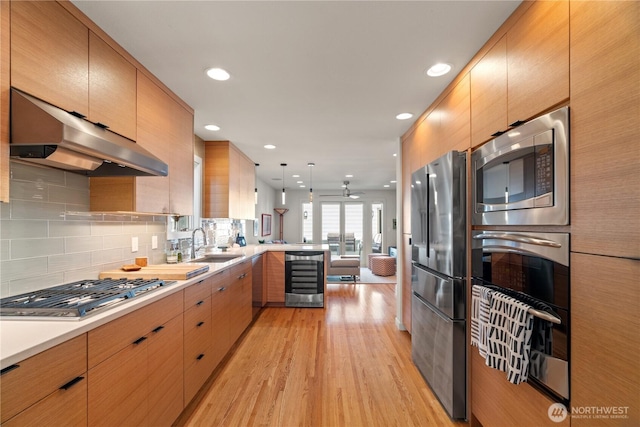 kitchen featuring a sink, a peninsula, under cabinet range hood, beverage cooler, and appliances with stainless steel finishes