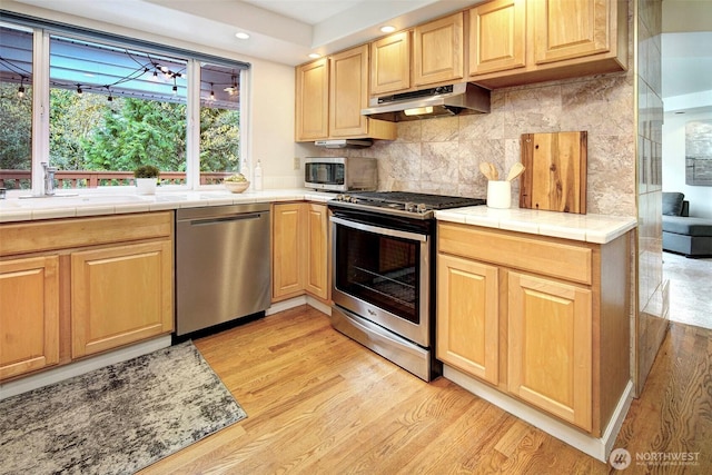 kitchen with a sink, stainless steel appliances, tile countertops, under cabinet range hood, and light wood finished floors