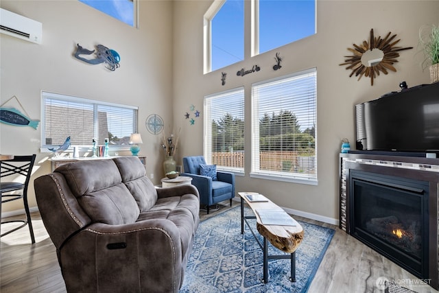 living area with a wall unit AC, wood finished floors, a glass covered fireplace, baseboards, and a high ceiling