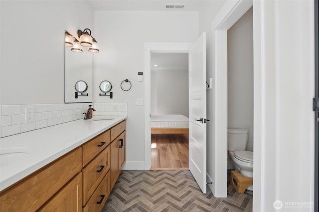 ensuite bathroom with toilet, brick patterned flooring, double vanity, baseboards, and decorative backsplash