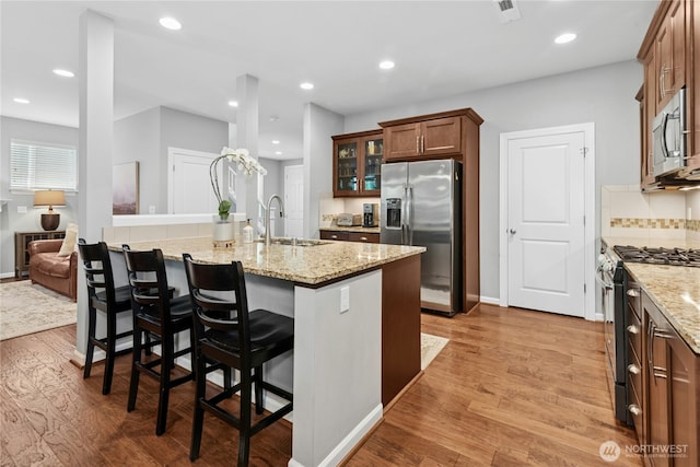 kitchen with appliances with stainless steel finishes, glass insert cabinets, light stone counters, light wood-style flooring, and a breakfast bar