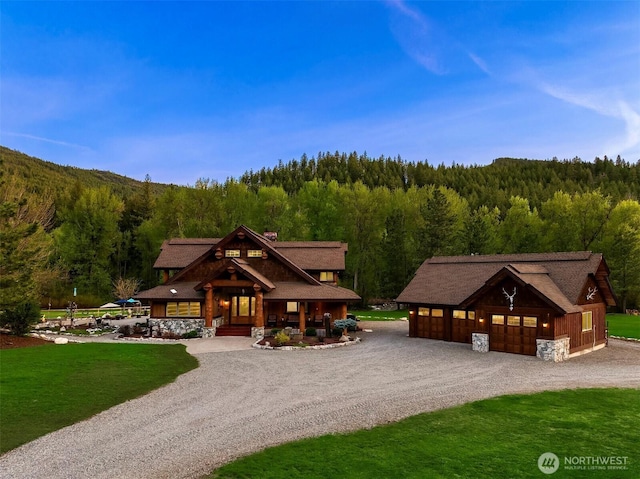cabin with a forest view, a porch, gravel driveway, a detached garage, and an outdoor structure