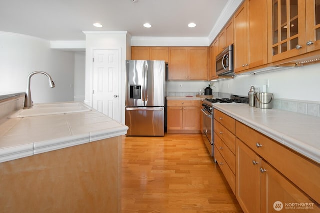 kitchen with stainless steel appliances, a sink, tile counters, crown molding, and light wood-type flooring
