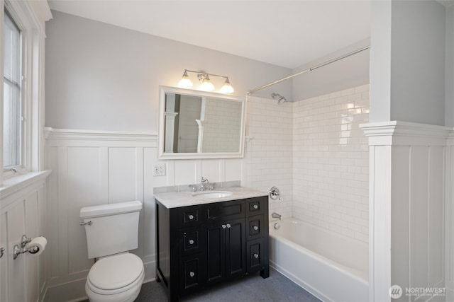 bathroom with toilet, a decorative wall, tub / shower combination, wainscoting, and vanity
