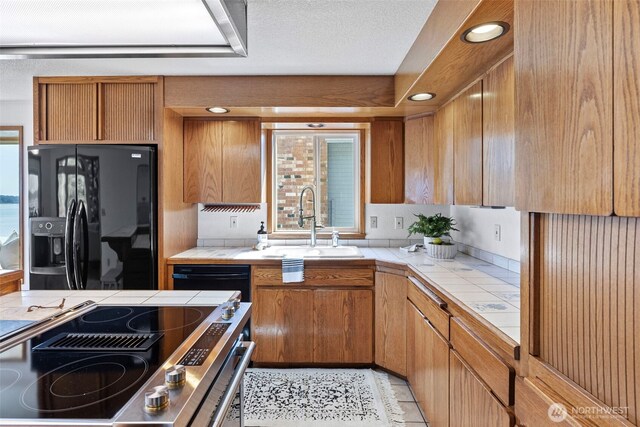 kitchen featuring tile countertops, black appliances, a sink, brown cabinetry, and recessed lighting