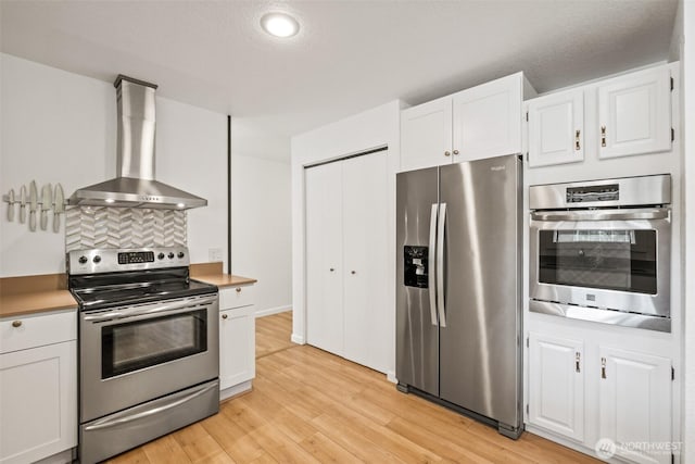 kitchen with stainless steel appliances, wall chimney range hood, light wood-style flooring, white cabinetry, and light countertops