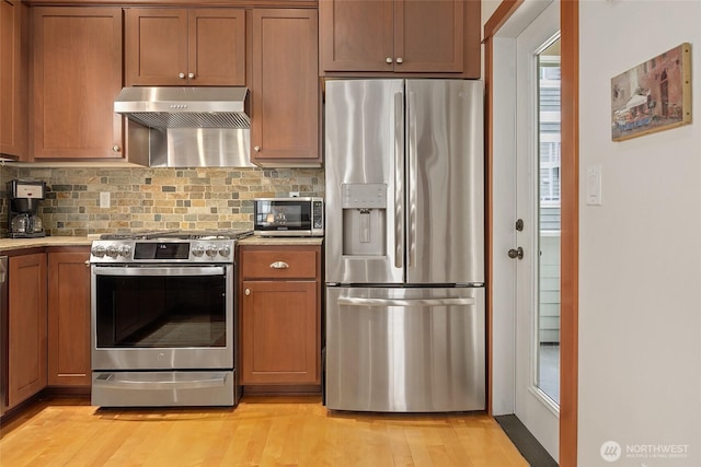 kitchen with appliances with stainless steel finishes, ventilation hood, decorative backsplash, light wood-style floors, and brown cabinets