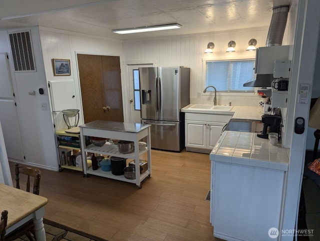 kitchen with stainless steel refrigerator with ice dispenser, tile countertops, a sink, light wood-style flooring, and wall chimney range hood