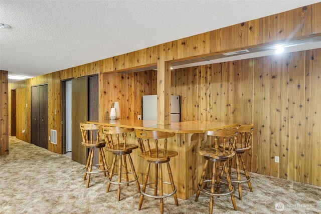 indoor dry bar with freestanding refrigerator, carpet, wood walls, and a textured ceiling