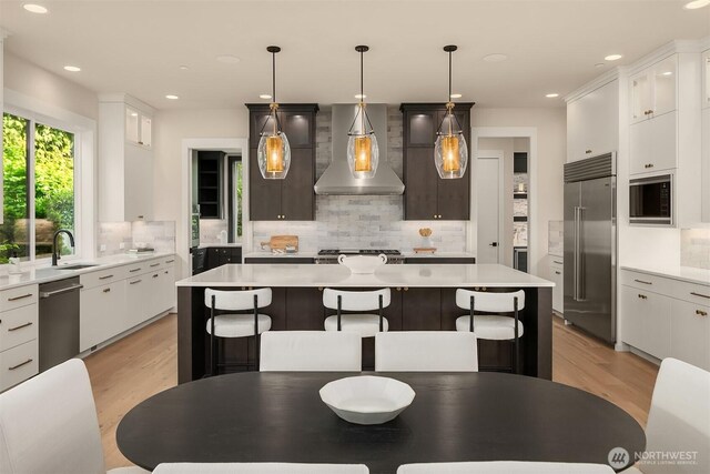 kitchen with wall chimney range hood, built in appliances, light wood-style floors, backsplash, and recessed lighting