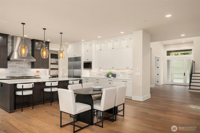 kitchen with wall chimney exhaust hood, built in appliances, a spacious island, light countertops, and tasteful backsplash