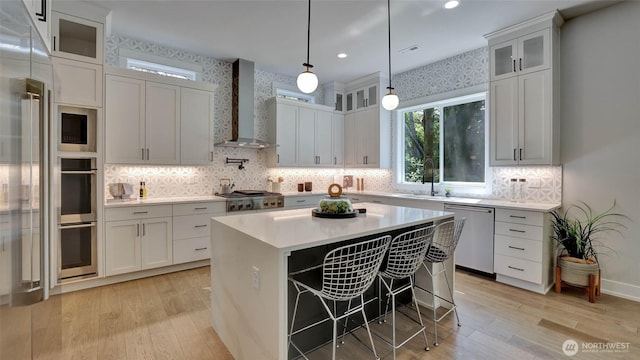 kitchen with wall chimney range hood, appliances with stainless steel finishes, a sink, light wood-style floors, and light countertops
