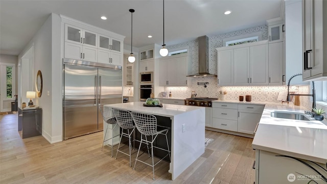 kitchen with built in appliances, wall chimney exhaust hood, a kitchen island, a kitchen bar, and a sink