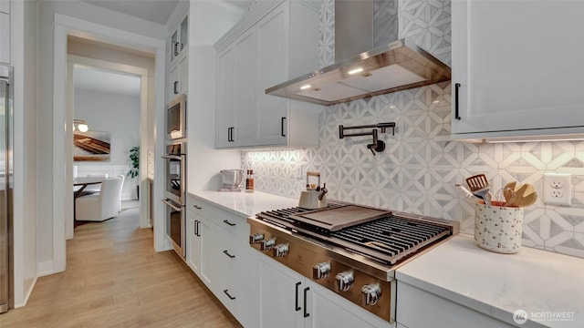 kitchen featuring wall chimney range hood, stainless steel appliances, light countertops, backsplash, and white cabinets