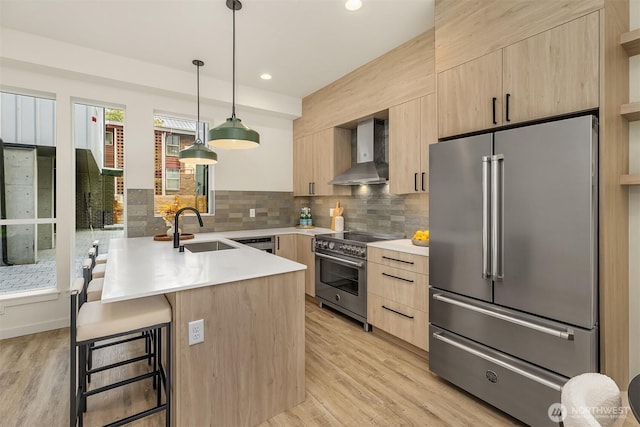 kitchen featuring high end appliances, light brown cabinetry, wall chimney range hood, modern cabinets, and a sink