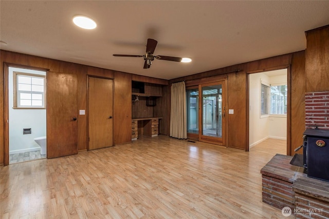 unfurnished living room featuring a wood stove, wood walls, light wood-type flooring, baseboards, and a ceiling fan