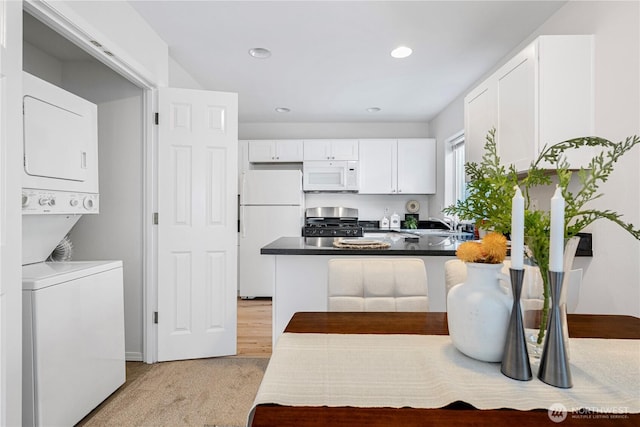 kitchen with white appliances, stacked washing machine and dryer, a peninsula, white cabinetry, and dark countertops