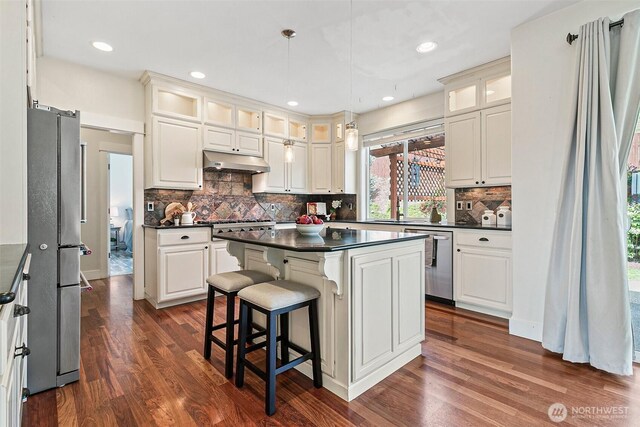 kitchen featuring stainless steel appliances, dark countertops, dark wood-style flooring, under cabinet range hood, and a center island