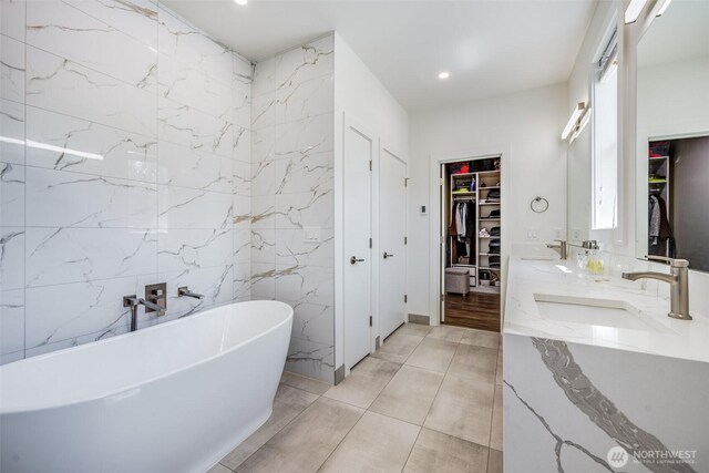 bathroom featuring a walk in closet, a freestanding bath, tile walls, double vanity, and recessed lighting