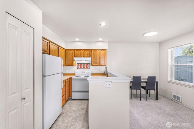 kitchen with white appliances, under cabinet range hood, a peninsula, brown cabinets, and light countertops