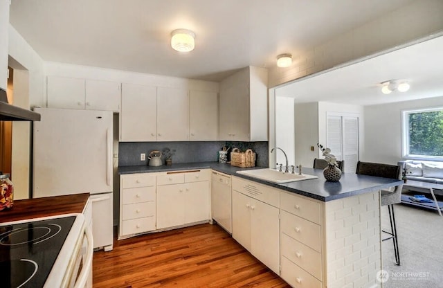 kitchen featuring a sink, white appliances, a peninsula, dark countertops, and a breakfast bar