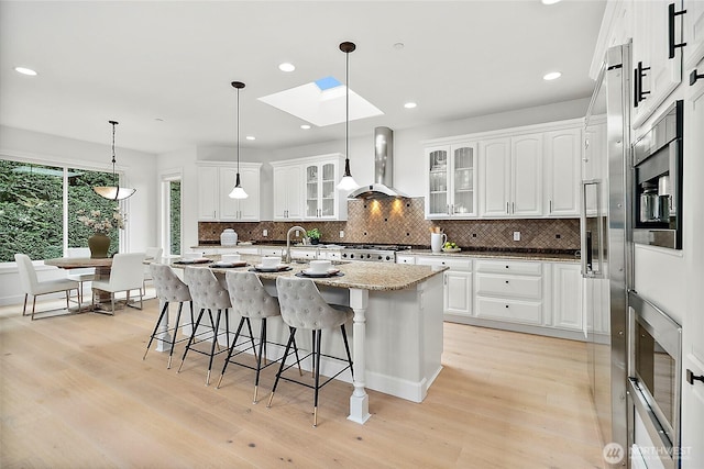 1038 Belfair Rd, Bellevue, WA 98004 - kitchen featuring wall chimney range hood, a skylight, a kitchen bar, backsplash, and light wood finished floors