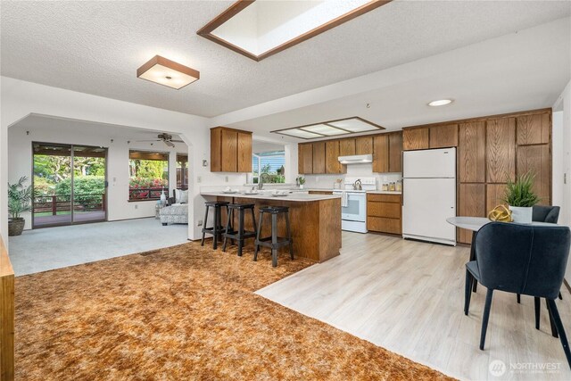 kitchen featuring white appliances, under cabinet range hood, brown cabinetry, open floor plan, and a peninsula