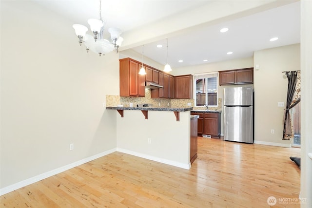 kitchen featuring freestanding refrigerator, under cabinet range hood, a peninsula, a breakfast bar, and light wood finished floors