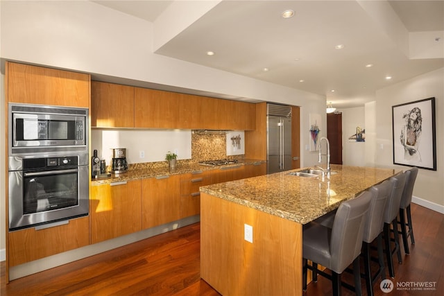 kitchen featuring built in appliances, a sink, brown cabinets, dark wood-style flooring, and a center island with sink