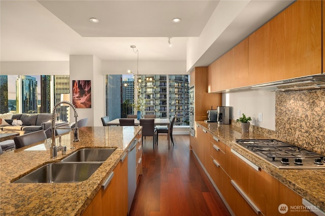 kitchen with a sink, stainless steel appliances, brown cabinetry, dark wood-style floors, and light stone countertops