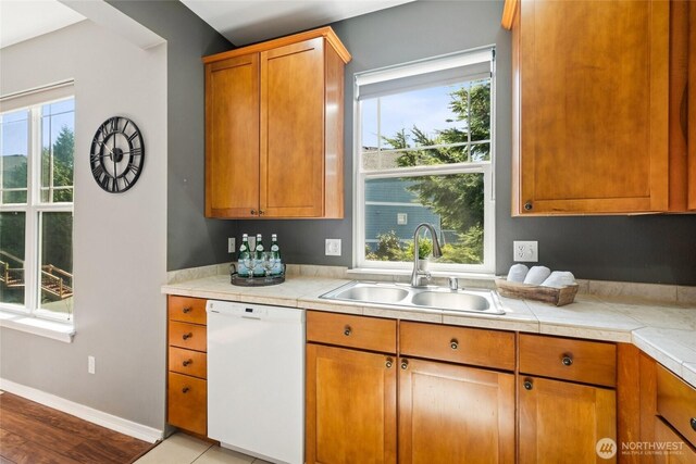 kitchen featuring white dishwasher, a sink, light countertops, and brown cabinetry