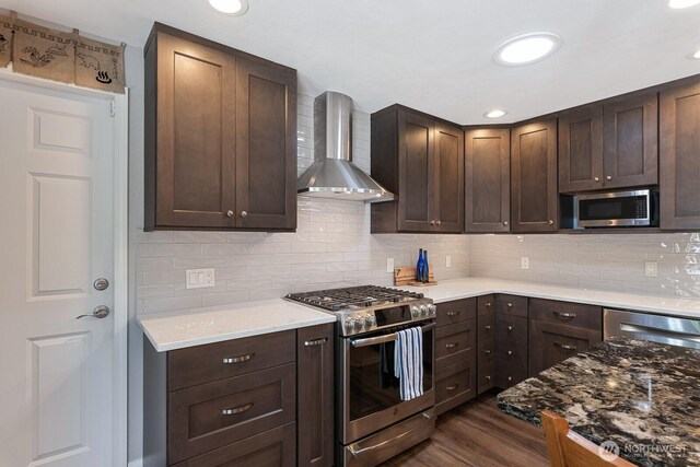kitchen with stainless steel appliances, wall chimney range hood, dark brown cabinets, dark wood finished floors, and decorative backsplash
