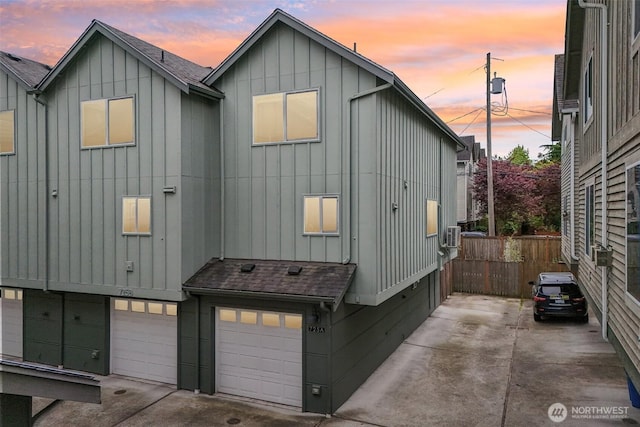 view of side of home with board and batten siding, an attached garage, a shingled roof, fence, and driveway