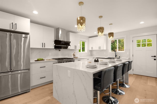 kitchen with stainless steel appliances, wall chimney range hood, light wood-style flooring, a sink, and recessed lighting