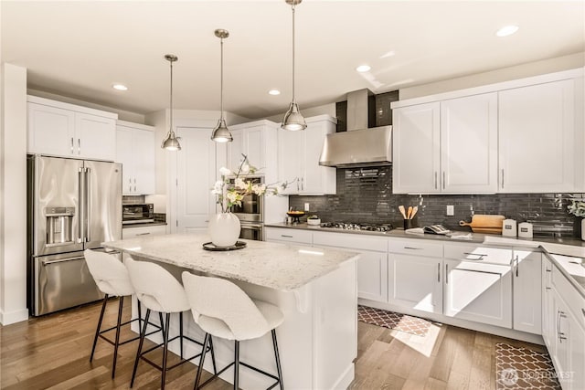 kitchen featuring appliances with stainless steel finishes, wall chimney exhaust hood, a kitchen island, wood finished floors, and decorative backsplash