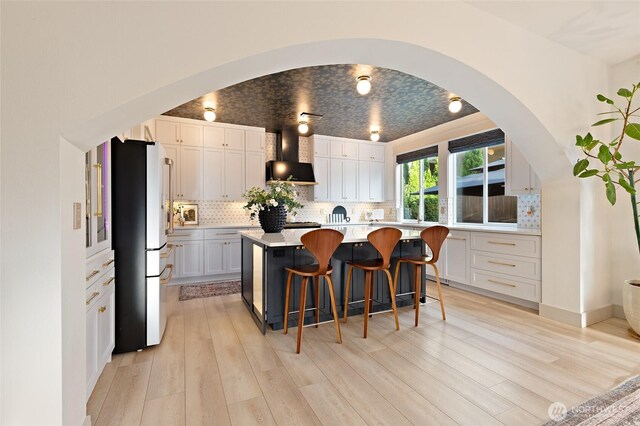 kitchen with freestanding refrigerator, wall chimney range hood, arched walkways, a breakfast bar area, and light wood-type flooring