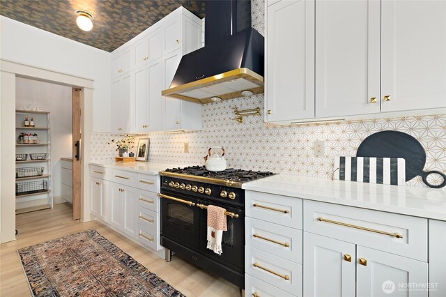 kitchen with range with two ovens, wall chimney exhaust hood, light wood-style flooring, backsplash, and white cabinets